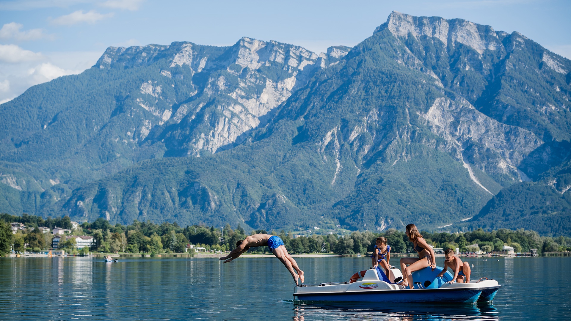 Ragazzo si tuffa da un pedalò sul lago, circondato dalla famiglia e dalle montagne.